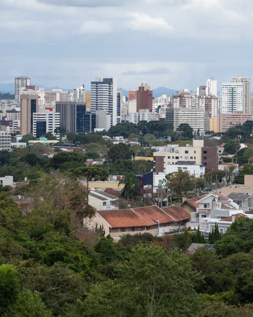 Vista de Curitiba, com área arborizada em primeiro plano e edifícios ao fundo